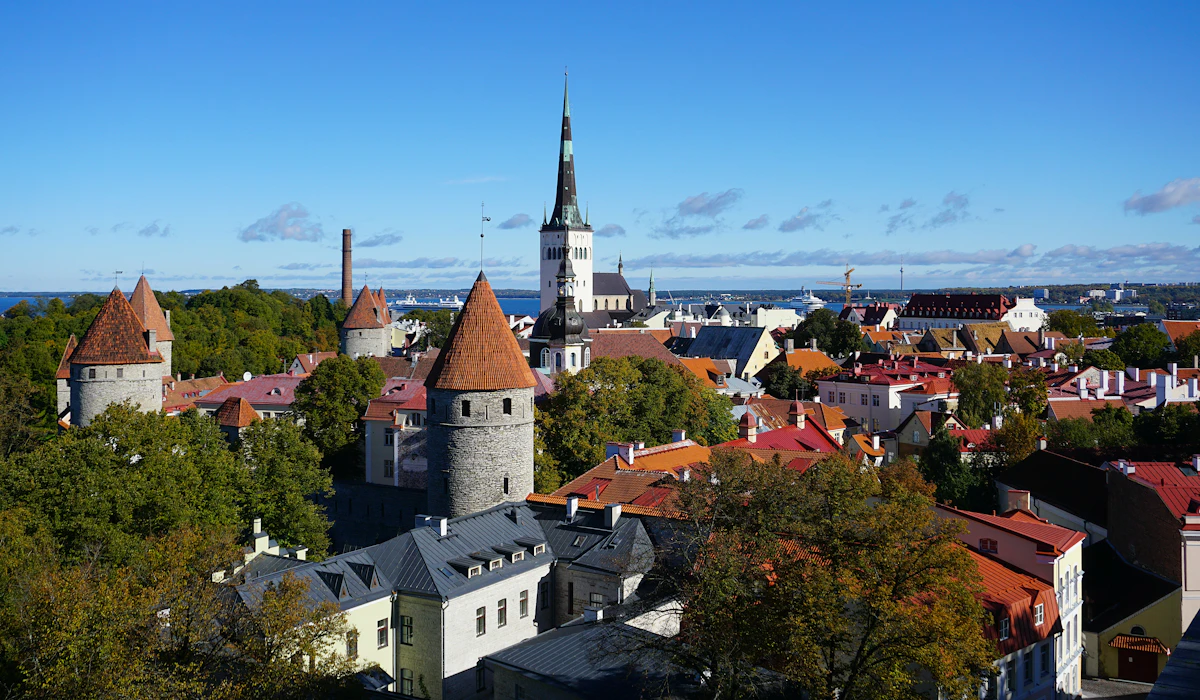 A view across the rooftops of Tallinn Old Town, with medieval wall towers under red conical roofs in the foreground and a tall church spire against the Baltic Sea beyond