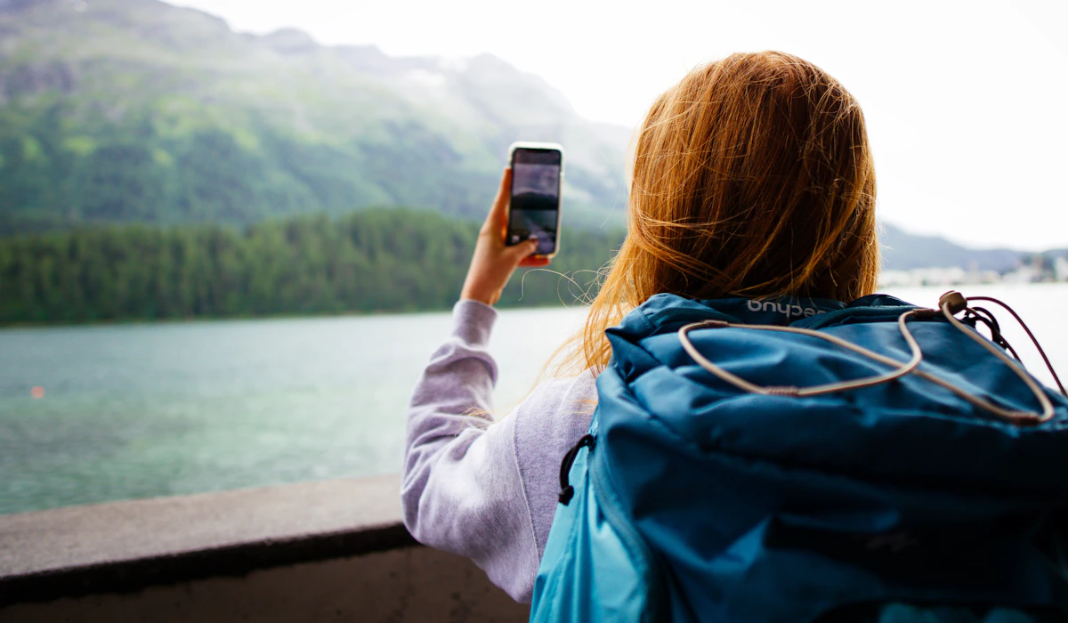 A traveler with a backpack holding up a smartphone toward a mountain lake view